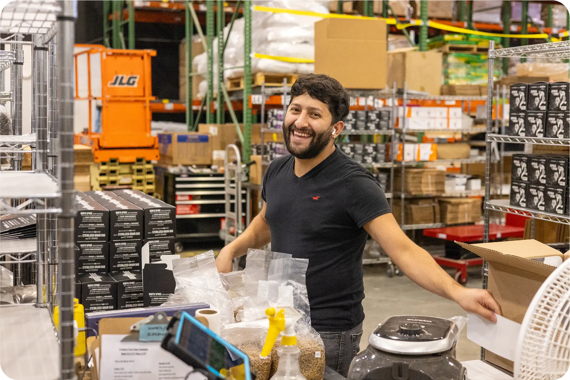 A smiling man with a beard stands in a warehouse among shelves and boxes, wearing a black t-shirt and handling packaged goods.