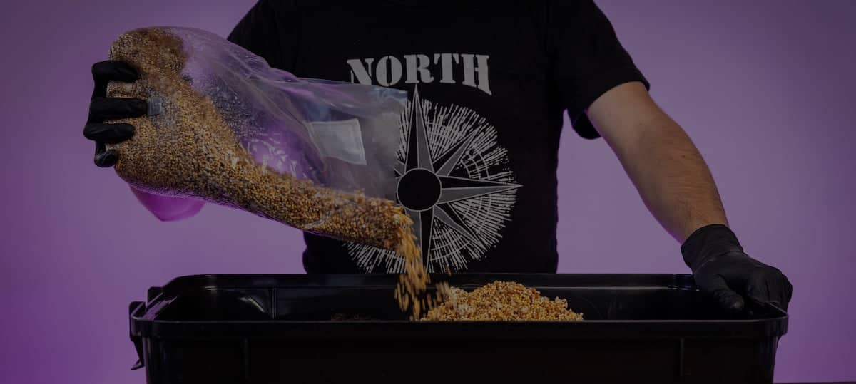 A person wearing black gloves pours grains from a plastic bag into a black container. The person is wearing a black shirt with NORTH written on it.