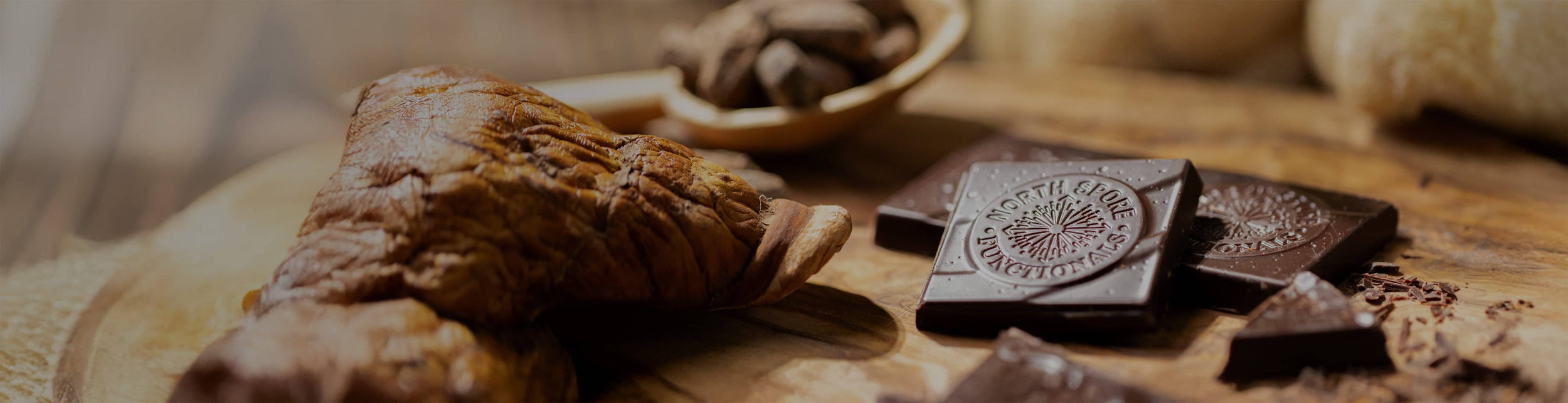 Close-up of dark chocolate squares with an embossed design on a wooden surface, next to a dried mushroom and a wooden spoon holding cacao beans.