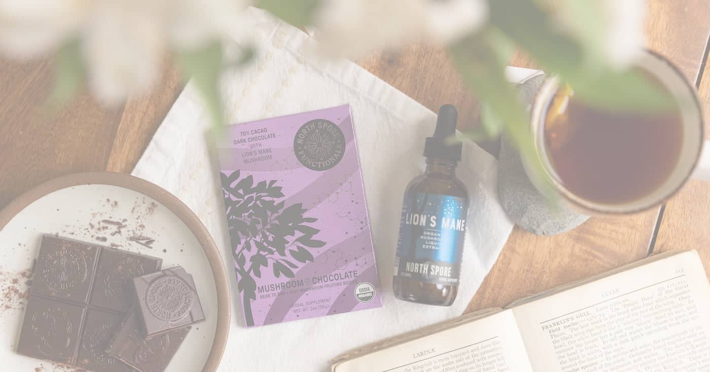 A flat lay of a chocolate bar, mushroom chocolate packaging, a Lions Mane supplement dropper, a cup of tea, and an open book on a wooden table.
