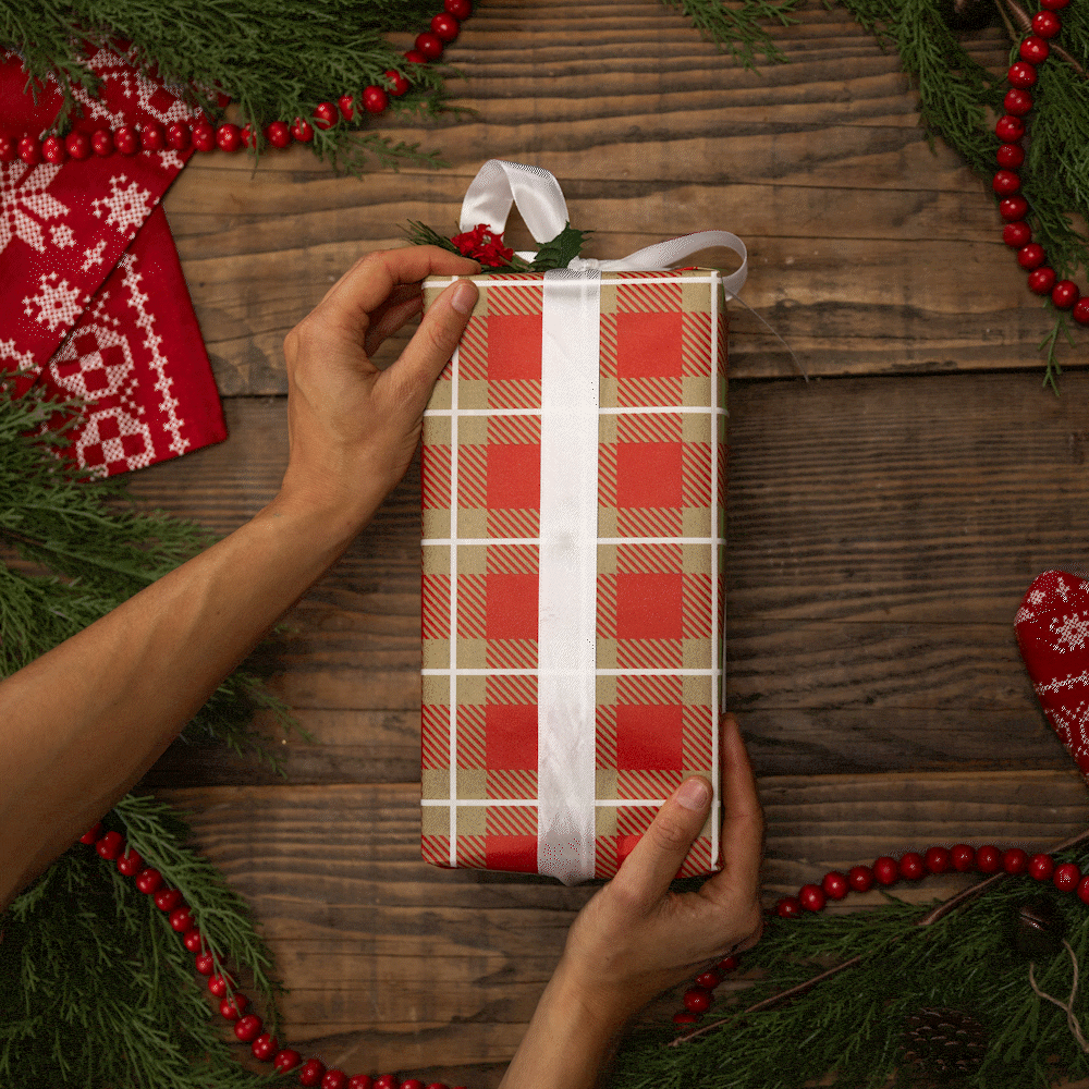 Hands holding a red and white plaid gift box with a white ribbon, surrounded by festive greenery, red beads, and a red patterned cloth on a wooden surface.