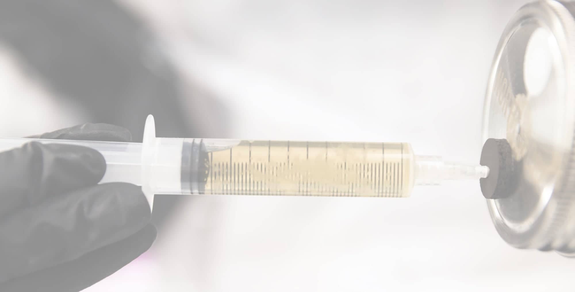 A gloved hand holds a syringe filled with a yellowish liquid, drawing it from a metal container against a blurred background.