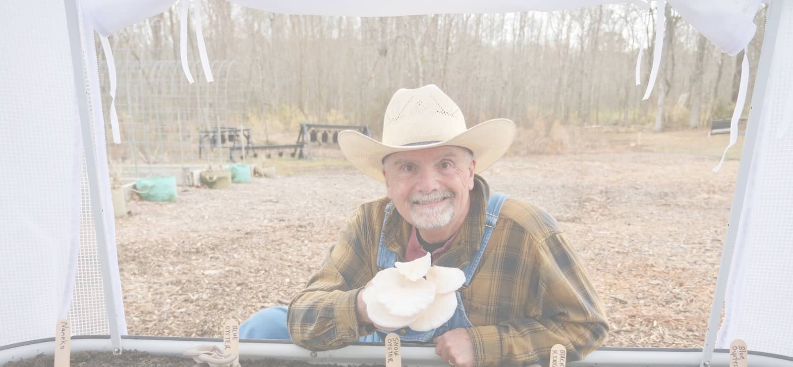 A smiling man in a cowboy hat and plaid shirt holds a cluster of mushrooms, viewed through a mesh enclosure in an outdoor, wooded area. Plant markers are visible in the foreground.