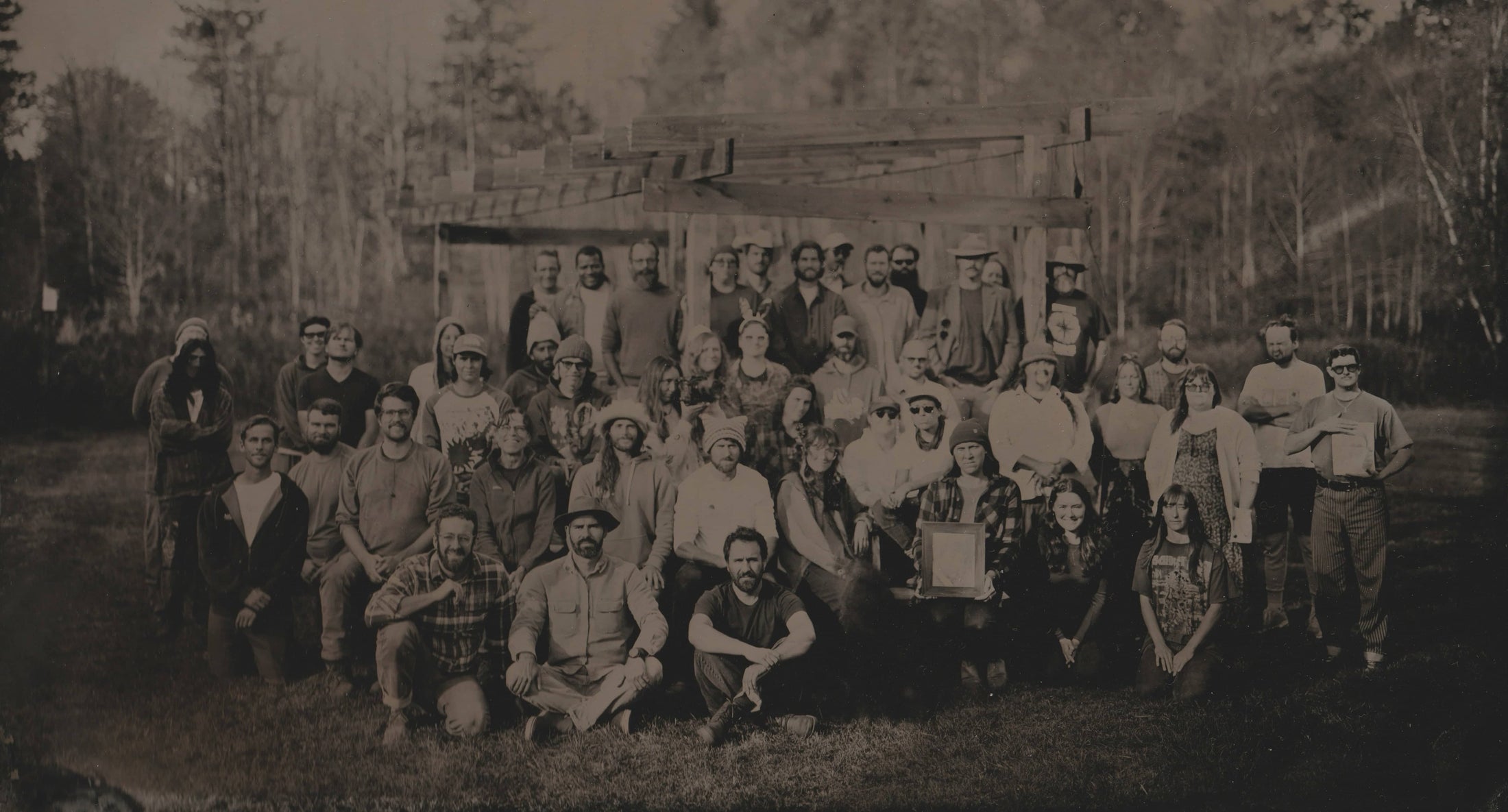 A large group of people, mixed genders, pose outdoors in front of a wooden structure, surrounded by trees; the photo has a vintage, sepia tone.