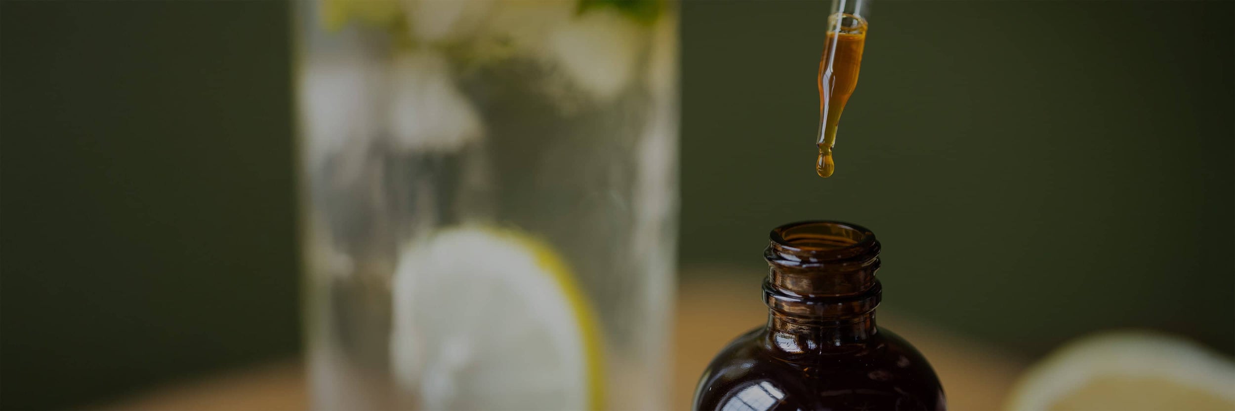 A dropper with brown liquid hovers over an open amber bottle; a glass of water with lemon slices is blurred in the background.
