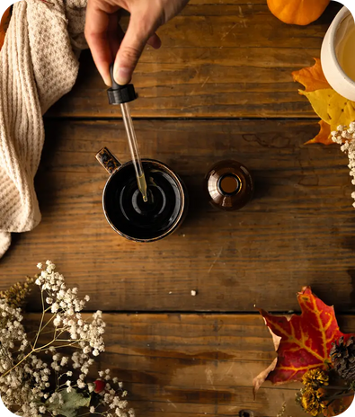 A hand holds a dropper over a mug on a wooden table, surrounded by autumn leaves, a bottle, a cozy knit cloth, and dried flowers.