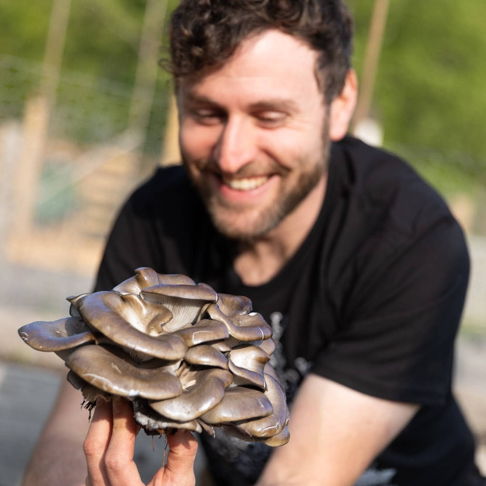 A smiling man with curly hair holds a large cluster of grayish-brown mushrooms outdoors, with a blurred green background.