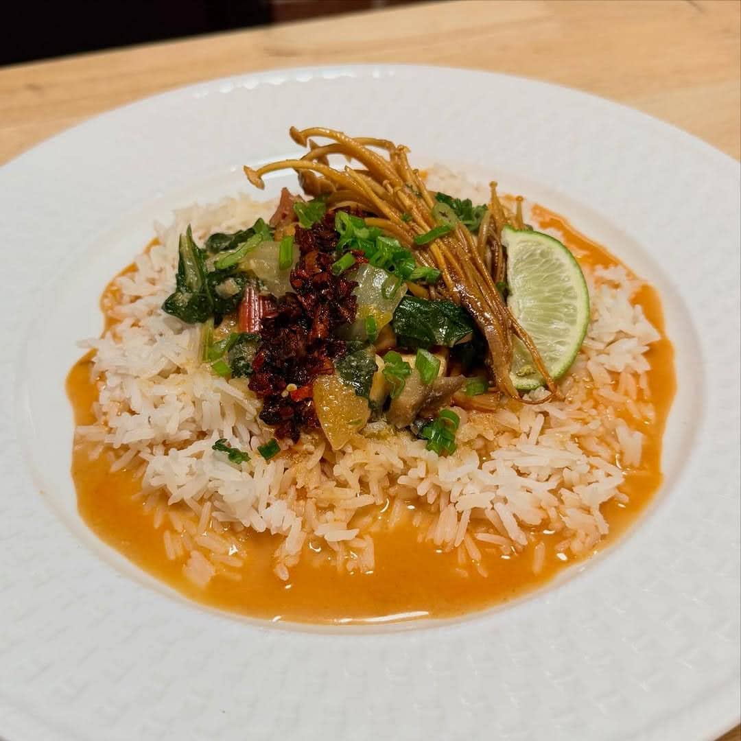 A plate of white rice topped with vegetables, greens, crispy mushrooms, a lime wedge, and garnished with herbs, served with an orange-colored sauce around the rice.