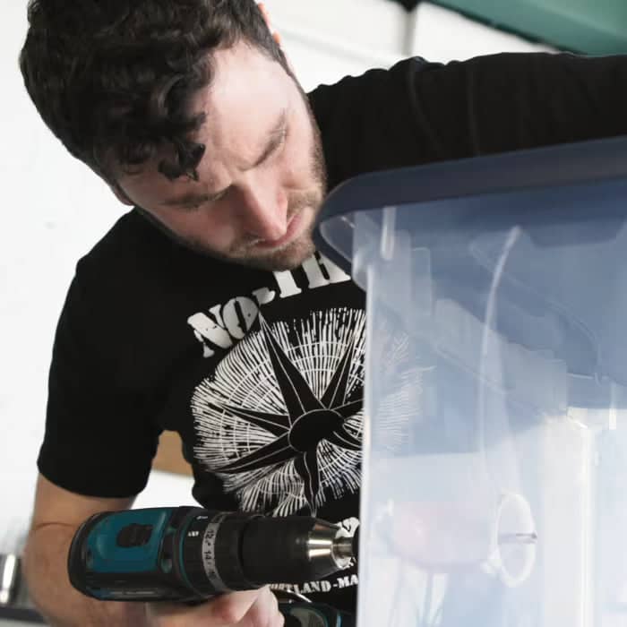 A man with curly hair uses a power drill on a clear plastic storage bin, focusing closely on his work.