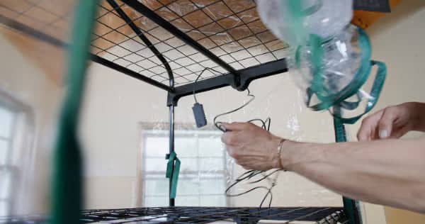A person’s hands are tying or adjusting green straps and wires to a black metal shelf frame indoors, with sunlight coming through a window in the background.