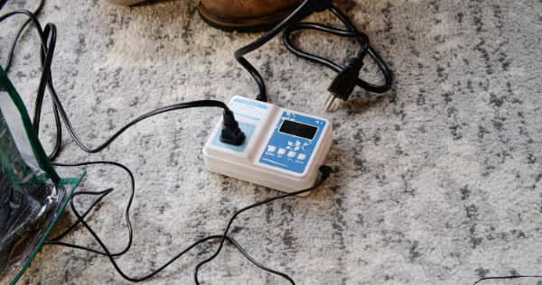 A white digital timer with multiple plugs and black cords on a light patterned carpet, with part of a brown shoe visible at the top.
