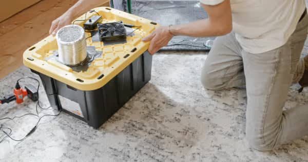 A person kneels on a rug, assembling a DIY air purifier with a fan and filter attached to a black and yellow plastic storage bin.