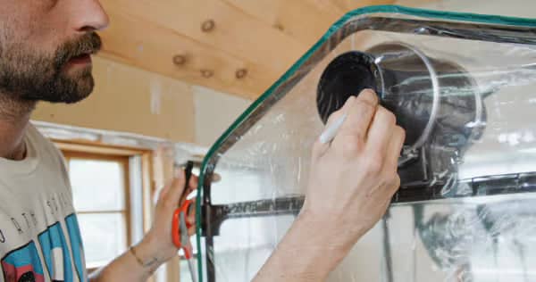 A person marks a circular hole on clear plastic sheeting attached to a metal frame, likely preparing for installation or construction work indoors.