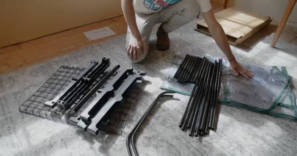 A person kneels on the floor, organizing metal rods, mesh panels, and parts, possibly assembling furniture or a metal structure on a carpeted surface.