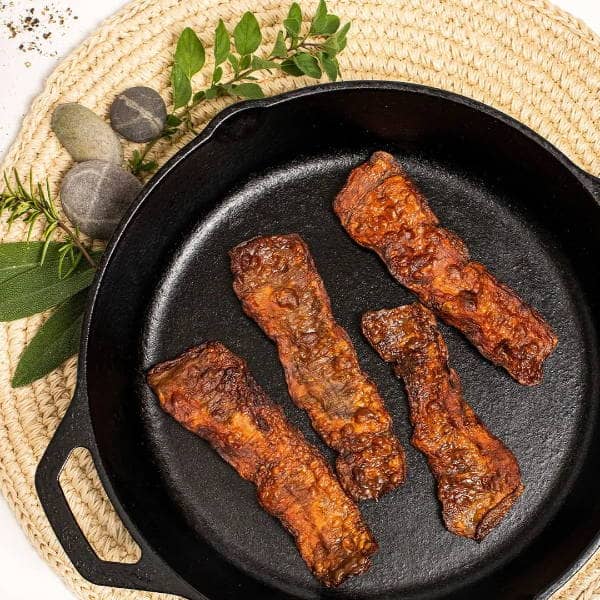 Four strips of cooked vegan bacon in a black cast iron skillet, placed on a woven mat with green herbs and decorative stones nearby.